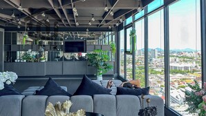 A modern living room with a large sofa, a coffee table, and a TV. There are plants and decorative items on the shelves. - The BASE Central Pattaya By INH Global (Pattaya)