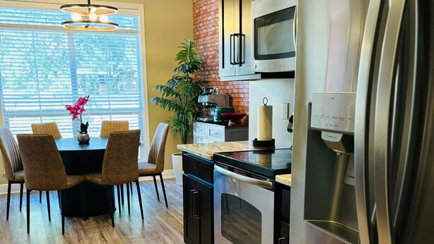 A modern kitchen with stainless steel appliances, a dining area with a table and chairs, and a brick accent wall. - Xquisite Nook, the ideal getaway in Daphne, AL! (Daphne)