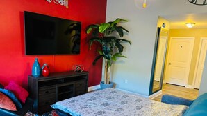 A living room with a red wall, a flat-screen TV, a potted plant, and a bed with a floral bedspread. - Xquisite Nook, the ideal getaway in Daphne, AL! (Daphne)
