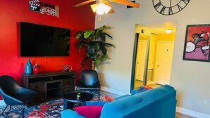 A living room with a red wall, a large flat-screen TV, a ceiling fan, a clock, and a door leading to another room. - Xquisite Nook, the ideal getaway in Daphne, AL! (Daphne)