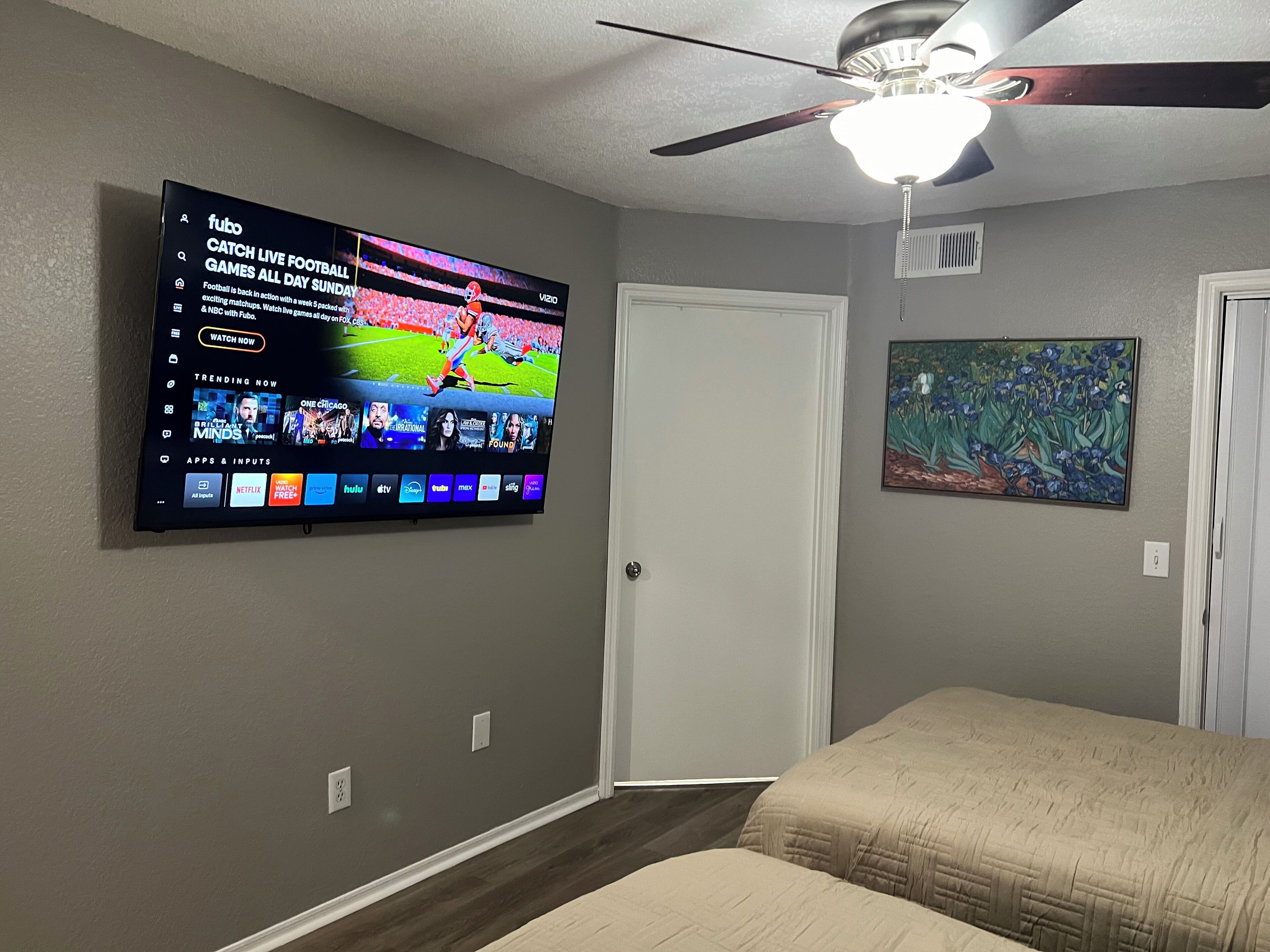 A bedroom with a TV mounted on the wall displaying a sports channel, a ceiling fan, a door, and a painting.