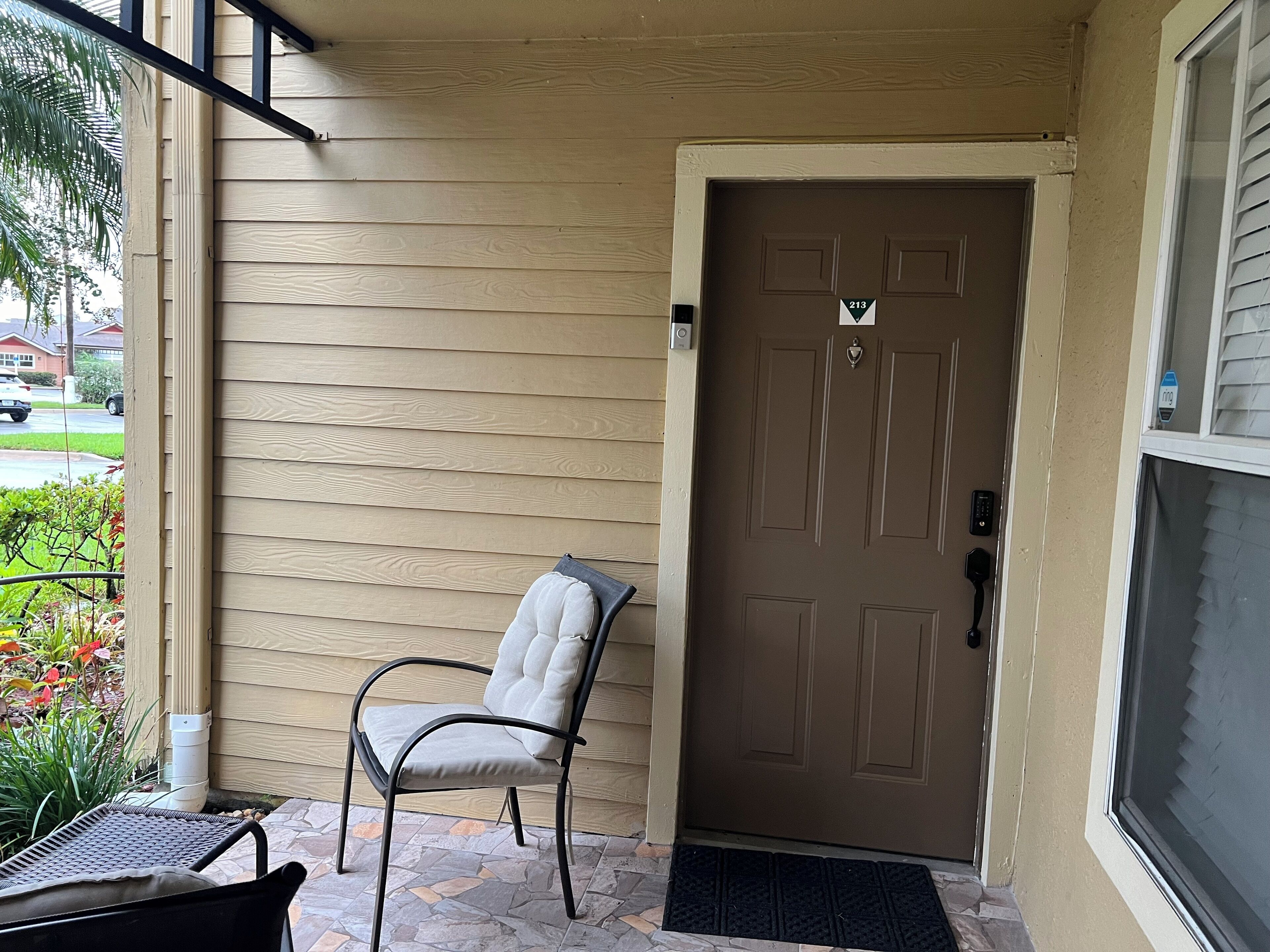Une porte en bois de couleur beige avec une plaque numérotée, une chaise en métal noir et un paillasson noir.