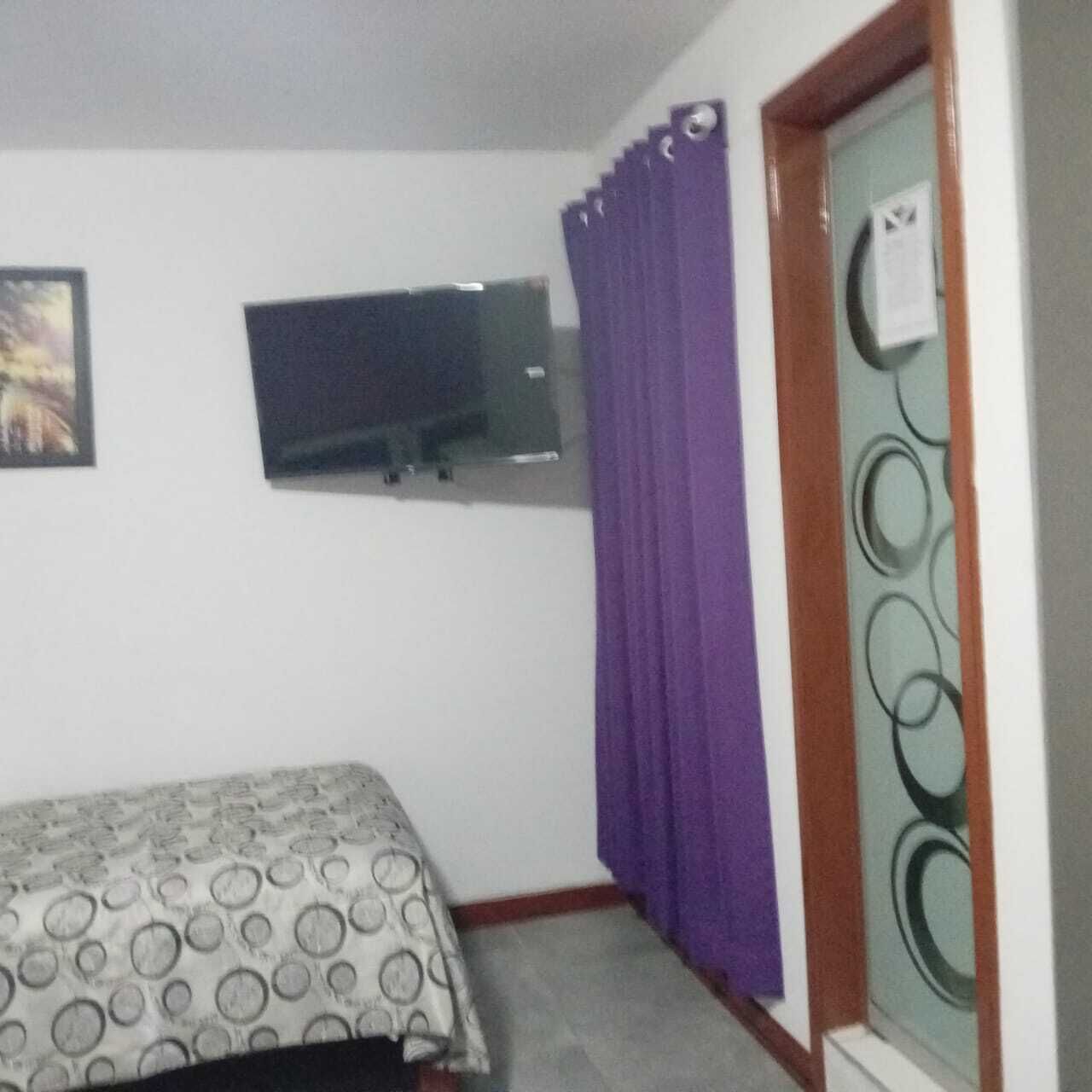 A room with a bed, a television mounted on the wall, and a window with purple curtains.