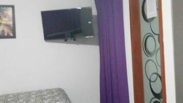 Una habitación con una cama, un televisor montado en la pared y una ventana con cortinas moradas.