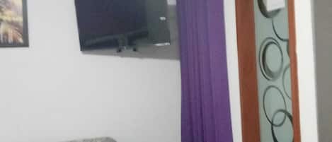 A room with a bed, a television mounted on the wall, and a window with purple curtains.