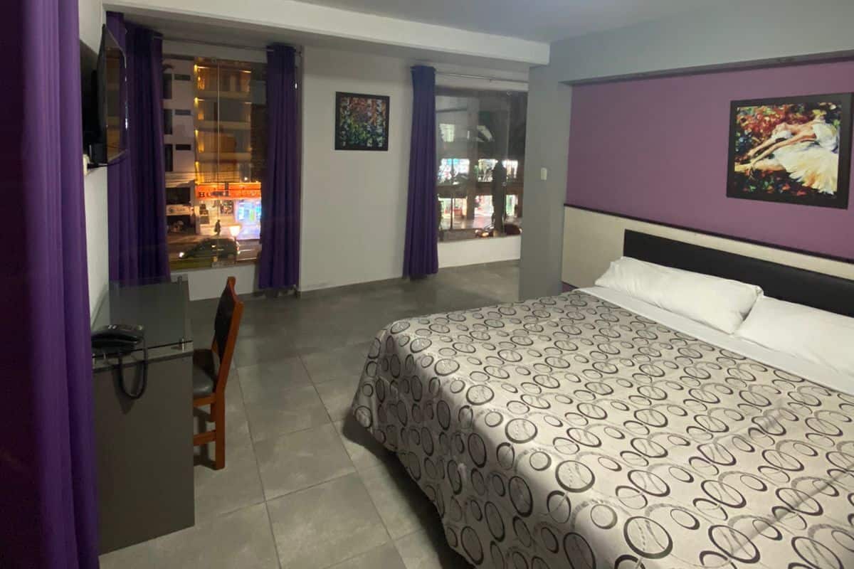 A hotel room with a bed, a desk, a chair, and a TV. There is a window with purple curtains, and a painting on the wall.