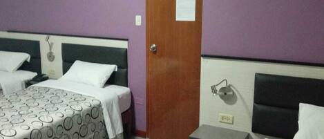 A hotel room with two beds, each with patterned bedding, a wooden door, and a small bedside table with a lamp.
