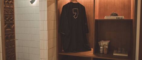 A compact bathroom with a toilet, a hanging robe, a suitcase, and a shelf with toiletries.
