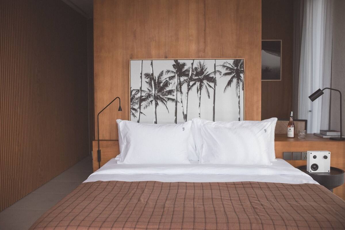 A hotel room with a bed, a wooden headboard, a wall-mounted picture of palm trees, a bedside table with a lamp, and a speaker.