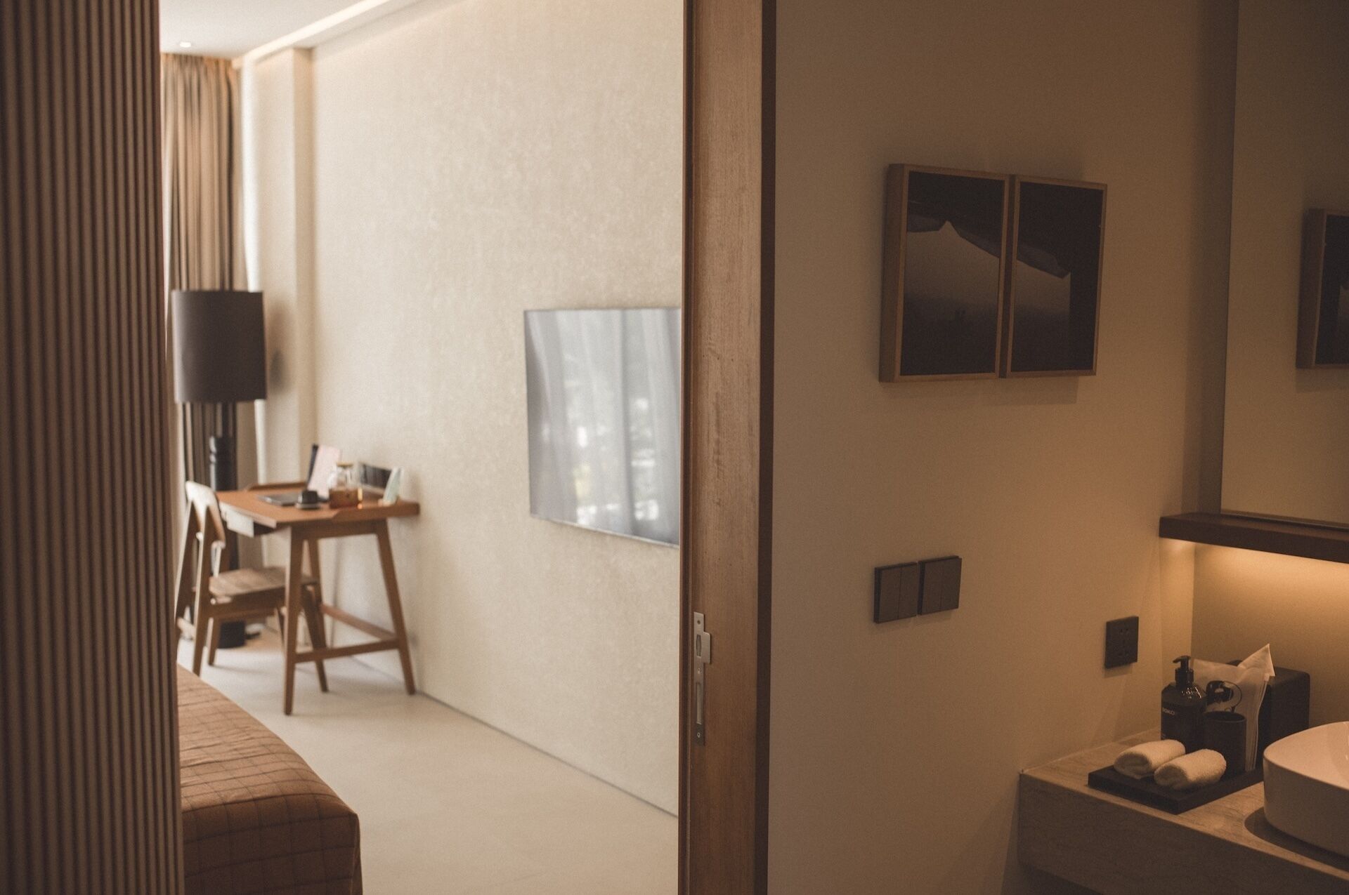 A hotel room with a wooden desk, a chair, a flat-screen TV, a wall-mounted artwork, and a bathroom with a sink and towel rack.