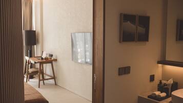 A hotel room with a wooden desk, a chair, a flat-screen TV, a wall-mounted artwork, and a bathroom with a sink and towel rack.