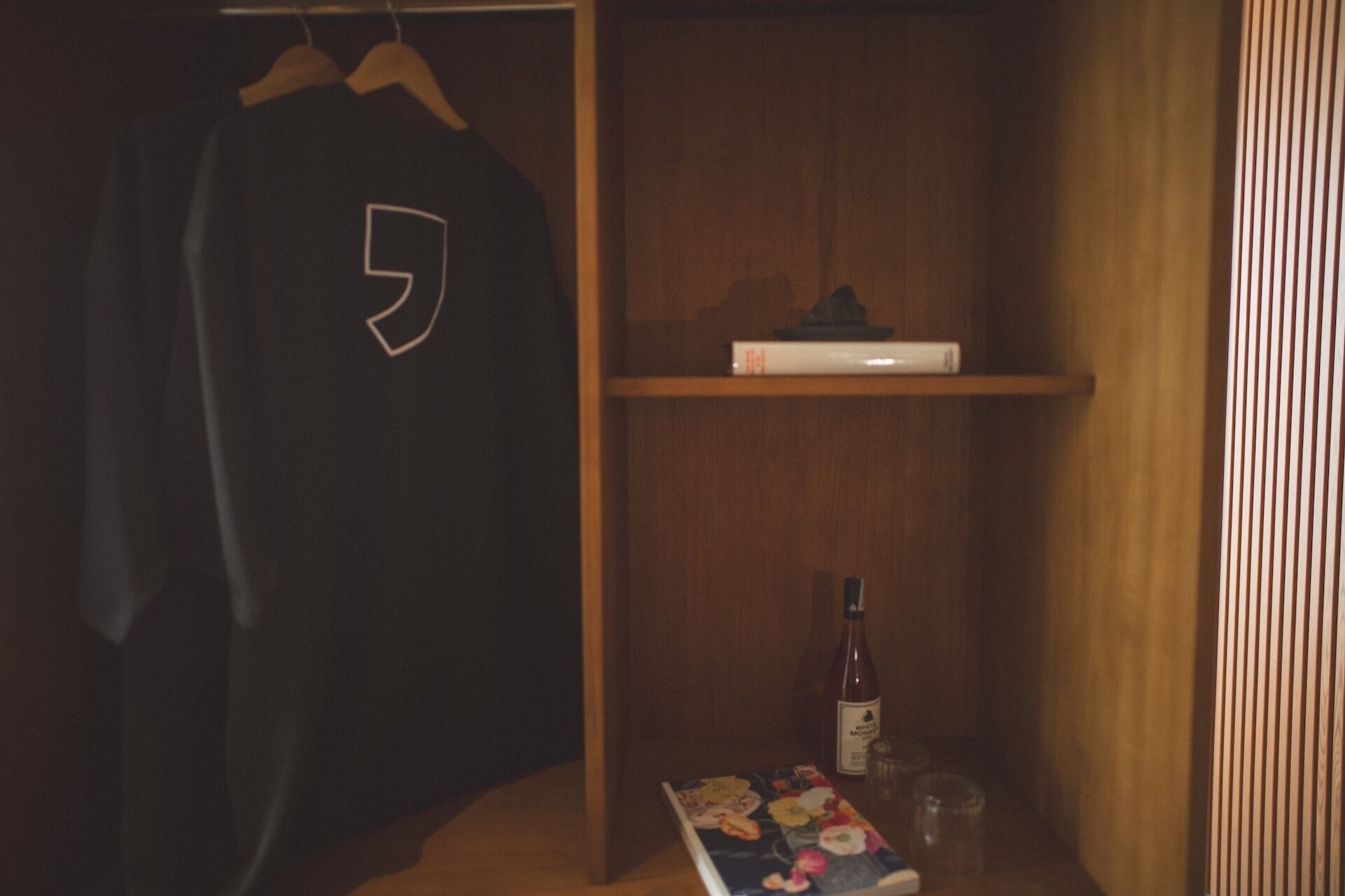 Una habitación con un armario de madera, una camiseta colgada, un estante con un libro y una botella.