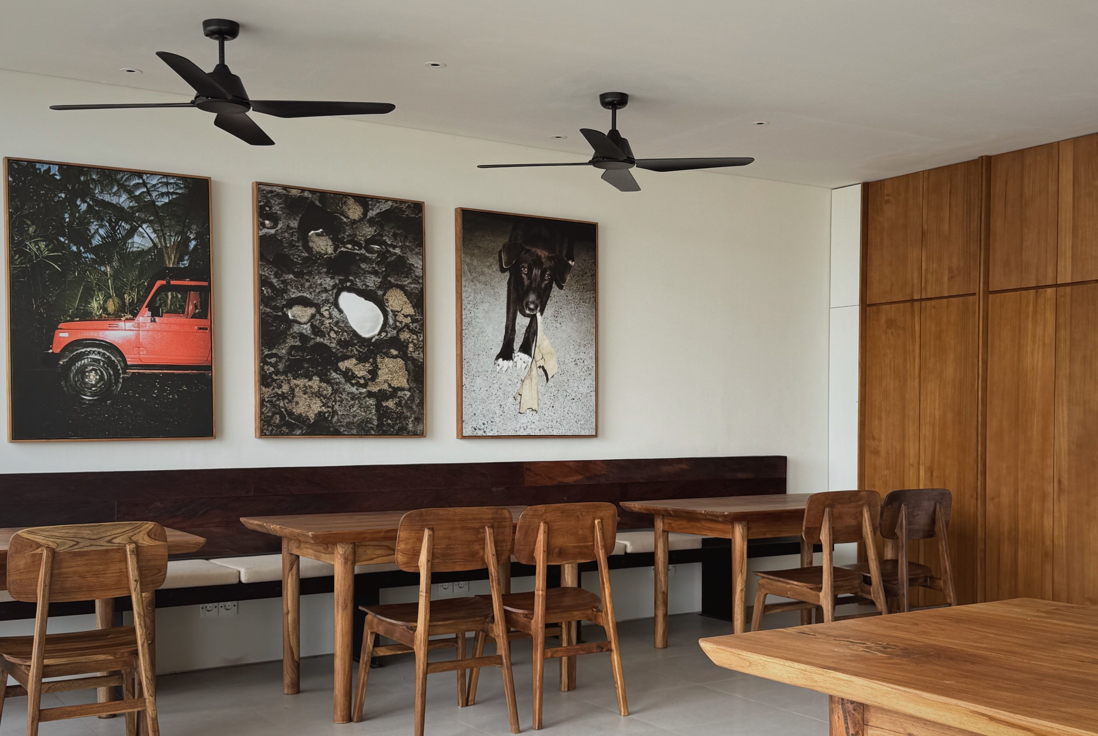 A dining area with wooden tables and chairs, ceiling fans, and three framed artworks on the wall.