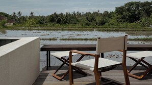 Een houten terras met een enkele klapstoel en een tafel, met uitzicht op een overstroomd gebied met weelderige begroeiing en palmbomen op de achtergrond.
