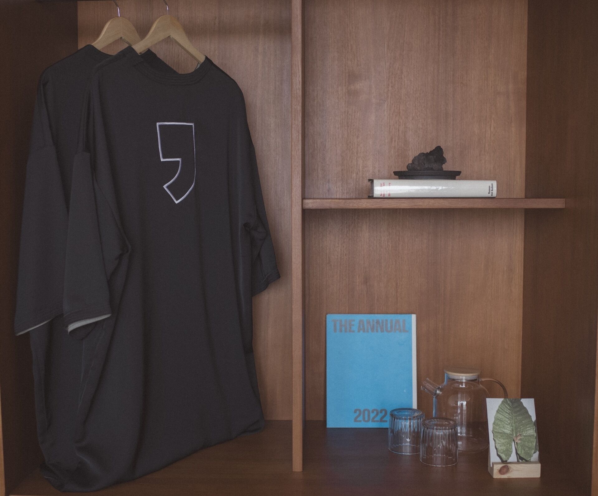 A black t-shirt with a white logo on a wooden hanger. Below, a blue sign reads "THE ANNUAL 2022" with a glass jar and a small plant beside it.