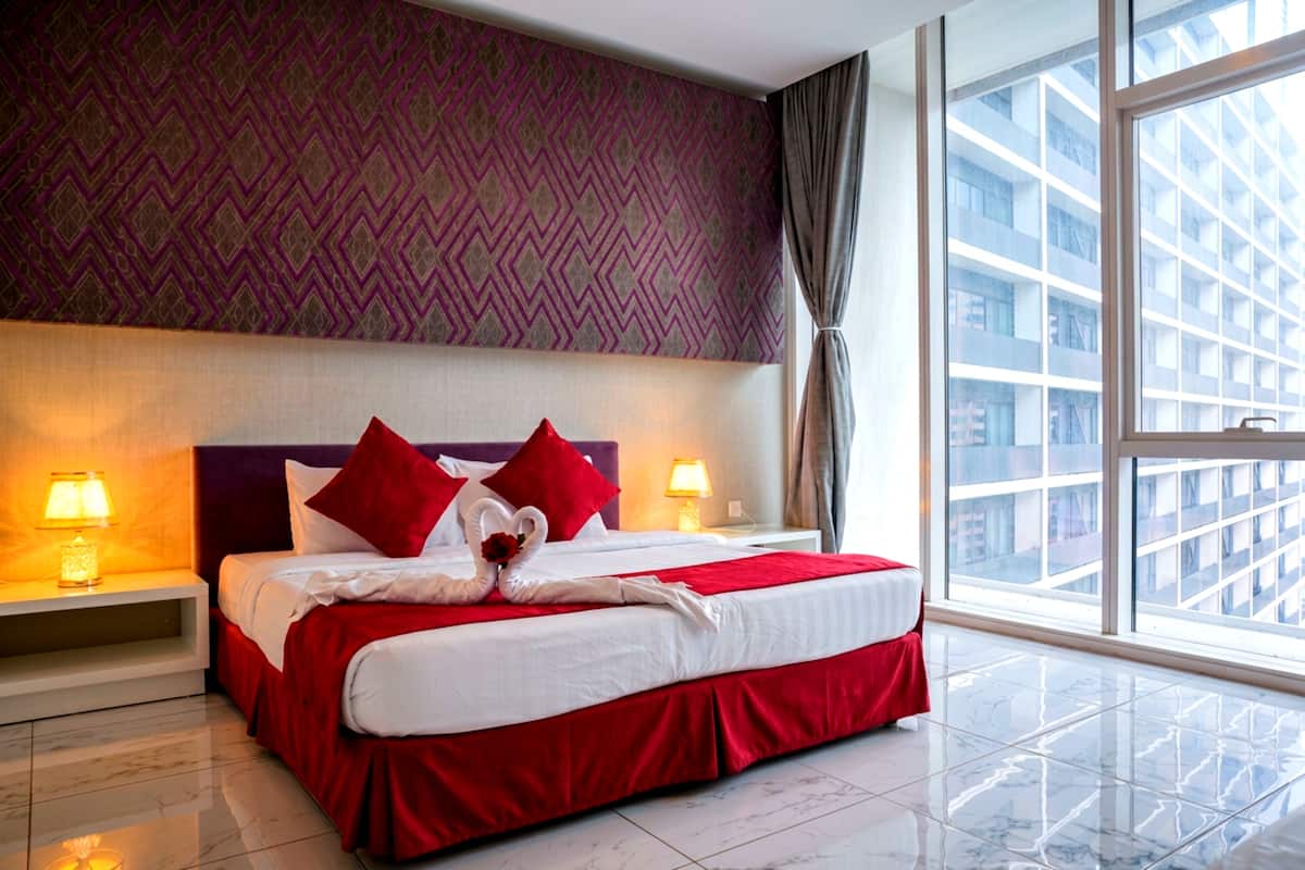 A hotel room with a large bed, red bedding, two bedside lamps, a patterned wall, and a view of buildings through a large window.