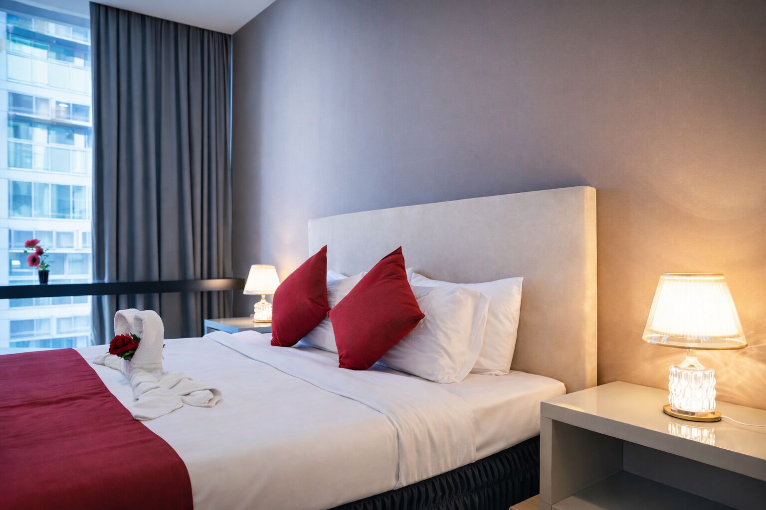 Camera d'albergo con un letto grande, cuscini rossi e bianchi, un comodino con una lampada e una finestra vista edifici.