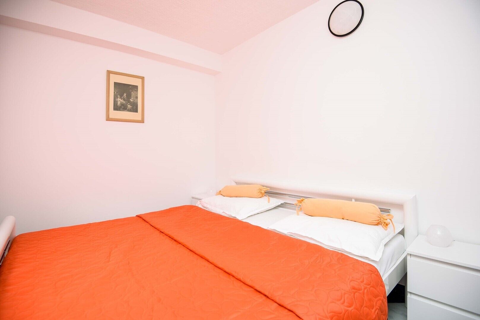 Une chambre avec un lit simple, des murs blancs, un tableau encadré accroché au mur et une table de chevet blanche.