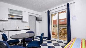 A compact kitchen with white cabinets, a black countertop, and a small TV. There is a dining area with a round table and blue chairs. The floor has a checkered pattern, and a colorful striped blanket is on the right. - Great 2+2 Apartment very close to the sea. (Kuciste)