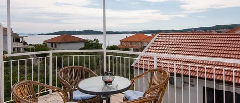 A balcony with wicker chairs and a table, overlooking a coastal area with buildings and a body of water.
