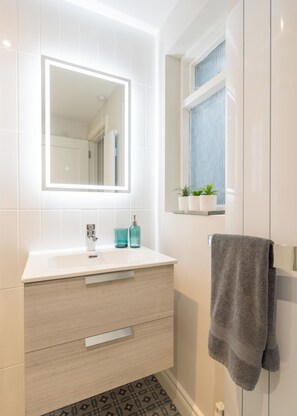 A modern bathroom with a white vanity, a mirror with integrated lighting, a towel rack, and a window with a view. - Cosy historic cottage in Hillsborough, near Belfast, Dublin & local attractions (Royal Hillsborough)