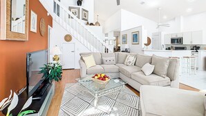A spacious living area with a large sofa, a glass coffee table with fruit, a TV, and a staircase leading to an upper level. - 🏰 Disney Area 3BR Pool Home | Family-Friendly | Minutes to Golf & Attractions (Davenport)