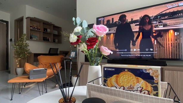 Ett modernt vardagsrum med en platt-TV, en bärbar dator på en stol, en vas med blommor och två burkar med mat på ett bord.