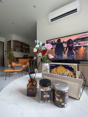 A modern living room with a flat-screen TV, a laptop on a chair, a vase of flowers, and two jars of food on a table. - LA Nest  (Ho Chi Minh City)
