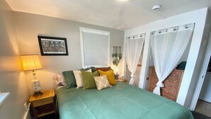 Un dormitorio con una colcha verde acolchada, varias almohadas, una lámpara de noche, un cuadro enmarcado en la pared y una ventana con cortinas blancas.