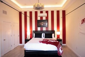 A hotel room with a bed, bedside tables, a wall-mounted air conditioner, a hanging light fixture, and a door. - Hoosier Suite Modern Studio Downtown on the Square (Bloomington)