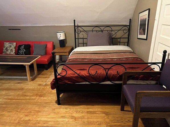 Een slaapkamer met een bed, een rode bank, een houten nachtkastje, een lamp en een stoel.