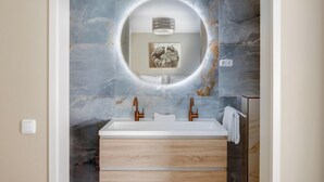 Ein modernes Badezimmer mit Marmorfliesenspiegel, einem runden Spiegel, einem weißen Waschbecken und Holzschränken.