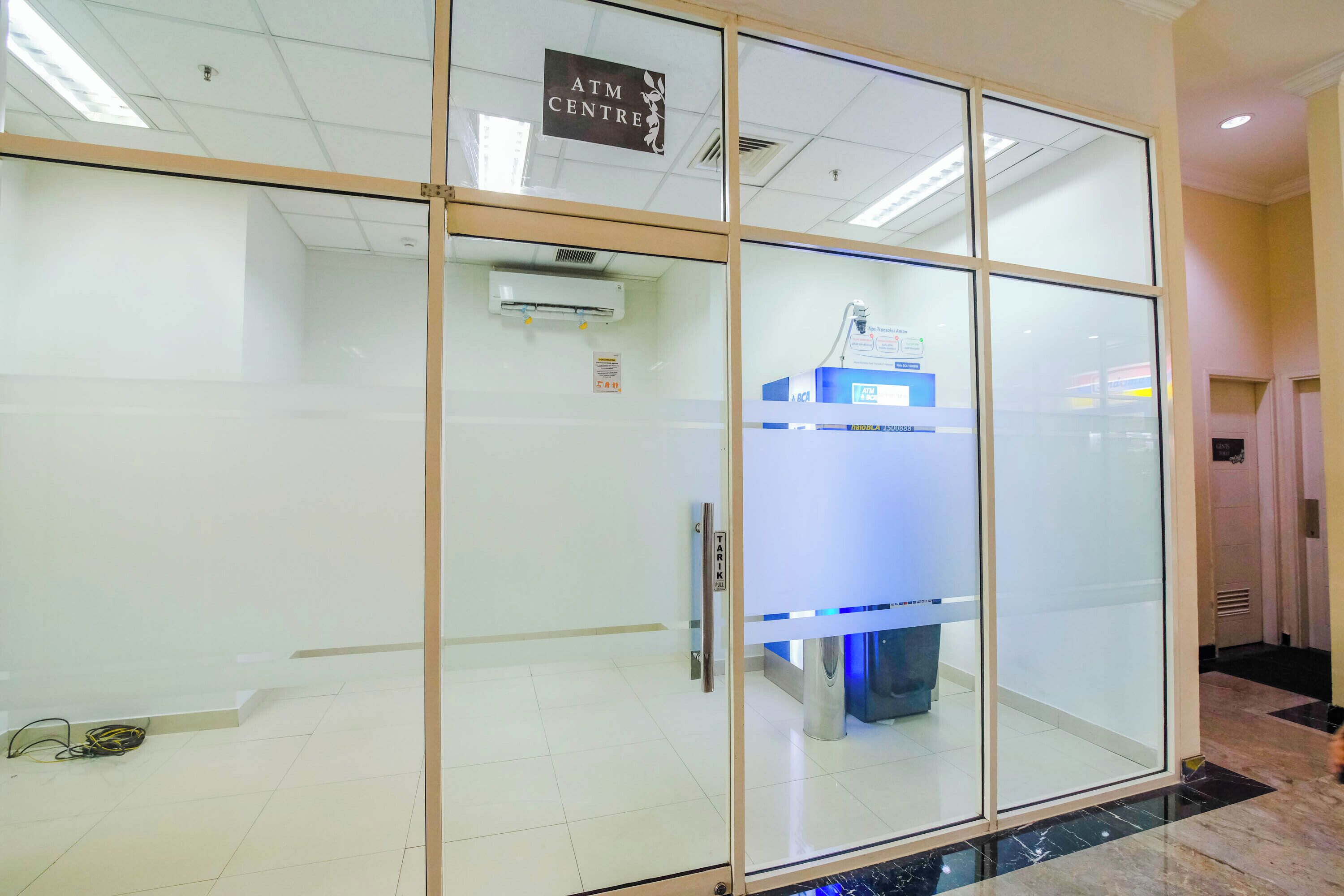A modern hotel lobby with a glass partition, an ATM, and a reception desk.