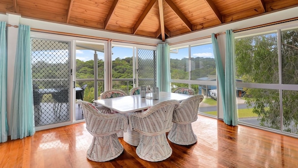 A dining area with a wooden table and wicker chairs, large windows with curtains, and a view of greenery and buildings. - Sundowner is the perfect escape for a relaxing holiday (Point Lookout)