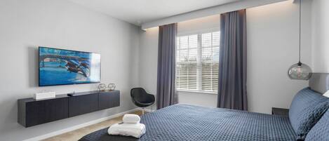 A modern bedroom with a large bed, a flat-screen TV mounted on the wall, a dark-colored console table, and a window with curtains.