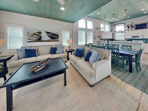 A spacious living area with a sofa and coffee table, a dining area with chairs and a table, and a kitchen with white cabinets and a large window.