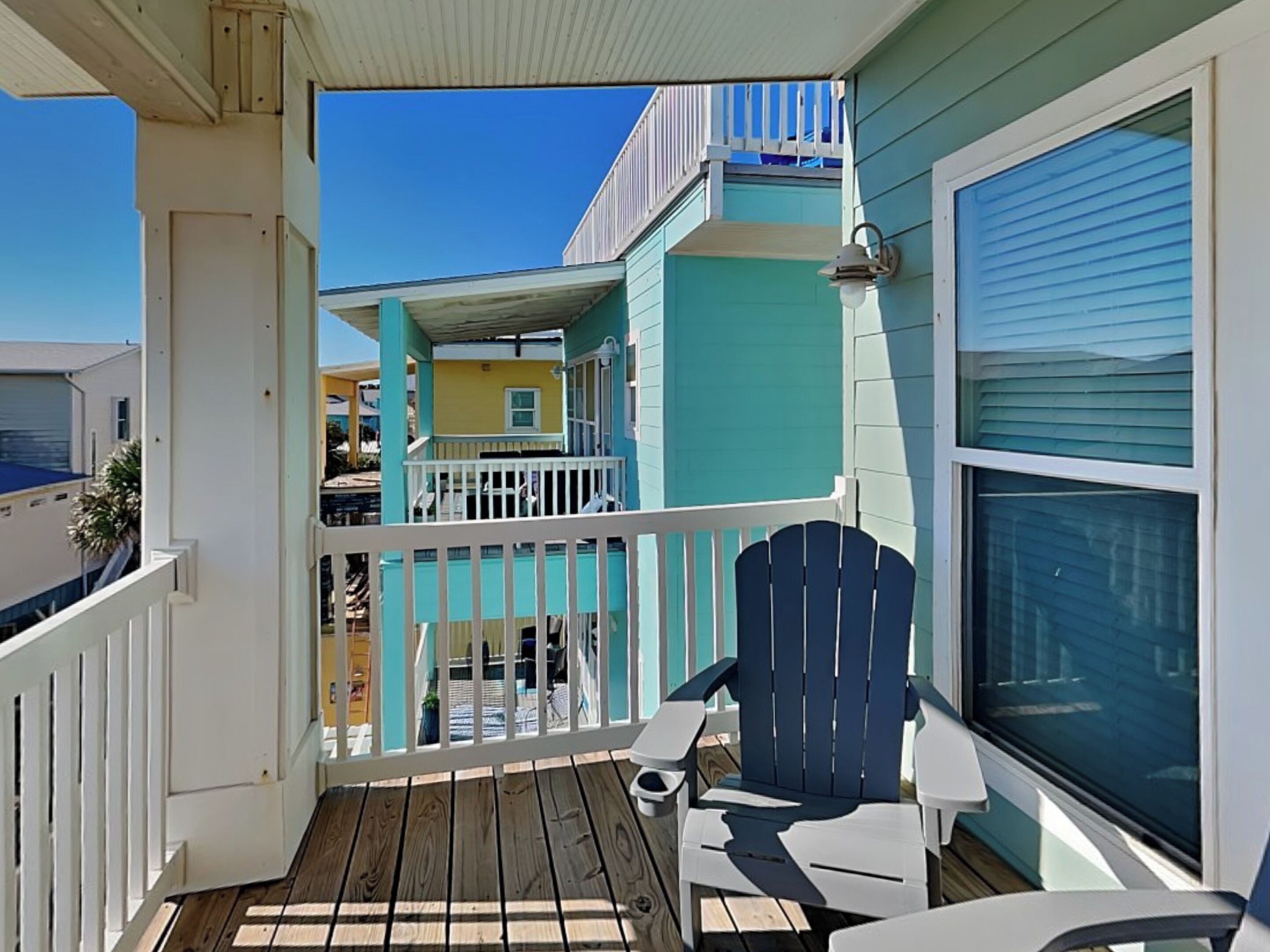 A balcony with a view of neighboring buildings, a white railing, and a blue Adirondack chair.