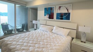 Un dormitorio con una cama grande, un ventilador de techo, dos mesitas de noche con lámparas, una ventana con vistas y un cuadro abstracto en la pared.