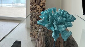 Un interior moderno con una mesa de vidrio que exhibe una escultura de coral azul, un jardín vertical de troncos con orquídeas rosadas y vistas a la playa a través de grandes ventanales.