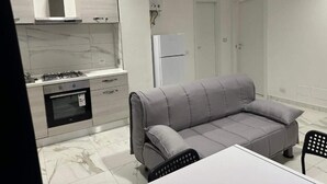 Moderni keittiö, jossa on kaasuliesi, uuni ja harmaa sohva. Keittiössä on myös valkoinen jääkaappi sekä ruokapöytä tuoleineen.