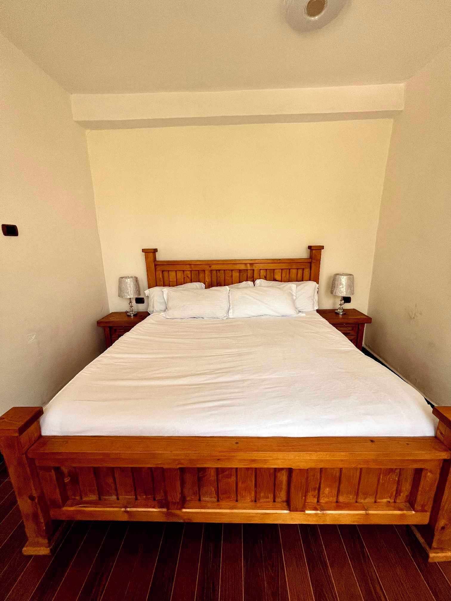 Een groot houten bed met witte lakens in een kamer met eenvoudige wanden en houten vloer.