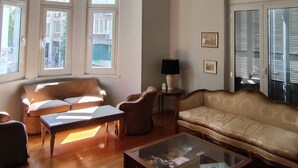 Un salon spacieux comprenant un canapé, des fauteuils, une table basse et une lampe. De grandes fenêtres laissent entrer la lumière naturelle et offrent une vue sur l’extérieur.