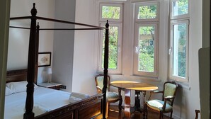 Un lit à baldaquin, une table ronde et une chaise dans une pièce dotée de grandes fenêtres et d’un lustre.