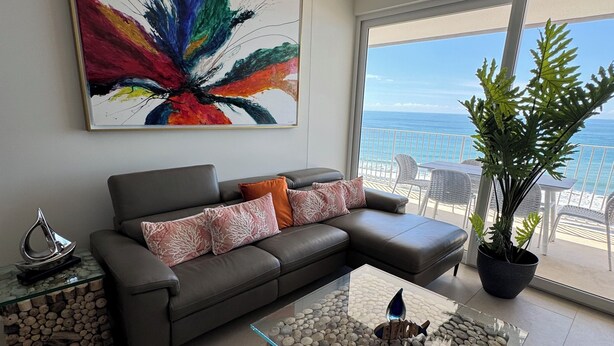 Ein modernes Wohnzimmer mit einem grauen Sofa, einem Glastisch mit Steinbasis, einem großen abstrakten Gemälde und Blick auf den Ozean durch große Fenster.