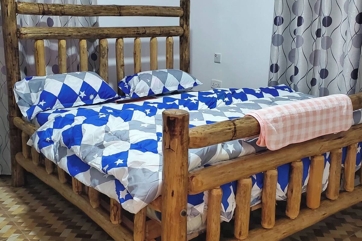 A wooden bed with a checkered bedspread and pillows, a wooden headboard, and a patterned curtain.