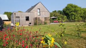 A modern wooden house with a sloped roof, a small garden with red and yellow flowers, and a fenced yard with red lounge chairs. - Contemporary home in a quiet location, a 10-minute walk from the beaches (Loctudy)