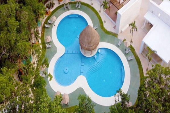 An aerial view of a resort with a large, uniquely shaped swimming pool surrounded by lounge chairs and a thatched-roof structure.