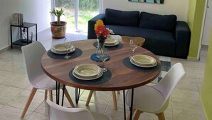 A dining area with a round table set for four, surrounded by white chairs. There is a ceiling fan above and a painting of a beach scene on the wall.