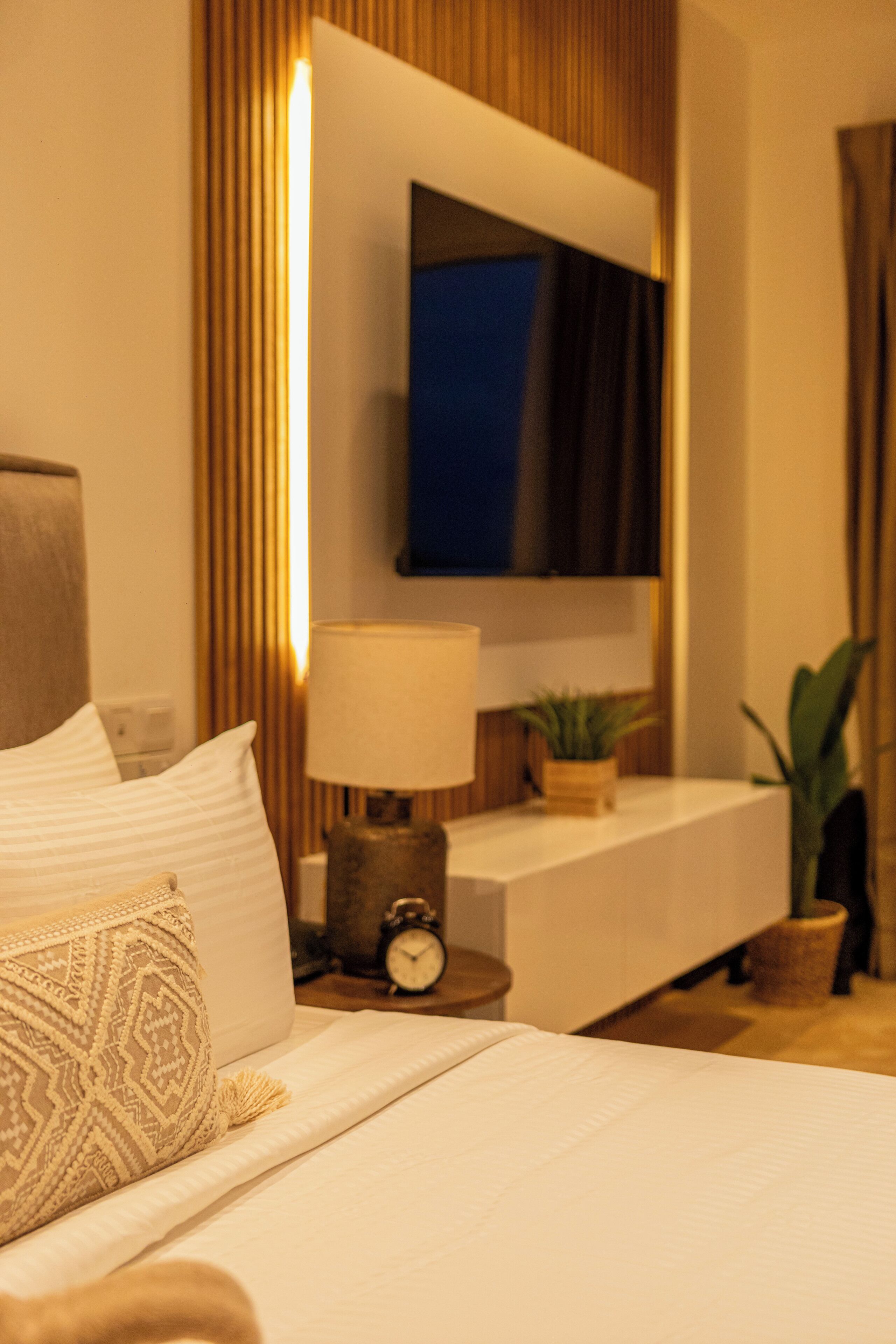 Una habitación de hotel con una cama, una mesita de noche con lámpara, un reloj, un televisor montado en la pared y una planta en maceta.