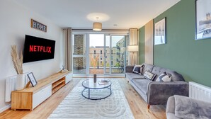A modern living room with a large TV displaying "NETFLIX," a sofa, a coffee table, and a view of buildings through large windows.